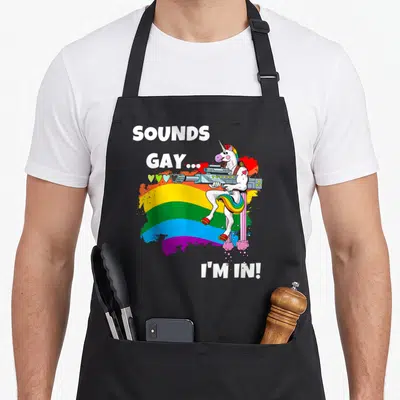 Sound unicorn gay pride apron - White unicorn holding gun on rainbow splatter with text SOUNDS GAY I M — shown on a black cotton canvas apron against a white background