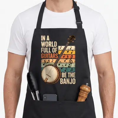 Guitar banjo apron - Retro banjo illustration with In A World Full Of Guitars Be The Banjo text and colorful stripes — shown on a black cotton canvas apron against a white background