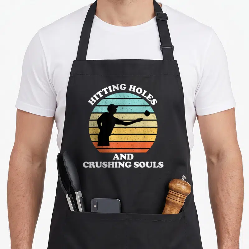 Hitting cornhole apron - Retro golfer silhouette in striped circle with white text HITTING HOLES AND CRUSHING SOULS — shown on a black cotton canvas apron against a white background