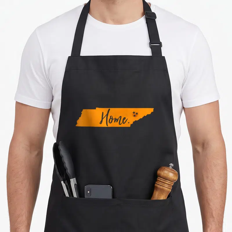 State football apron - Orange Tennessee map shape with grey Home script and three stars in a circle on the right side — shown on a black cotton canvas apron against a white background