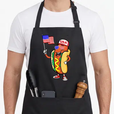Patriotic hot dog apron - Cartoon hot dog wearing USA cap and flag-patterned sunglasses holding American flag — shown on a black cotton canvas apron against a white background