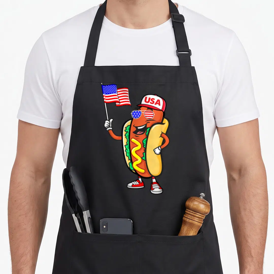 Patriotic hot dog apron - Cartoon hot dog wearing USA cap and flag-patterned sunglasses holding American flag — shown on a black cotton canvas apron against a white background