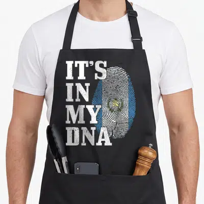 Dna guatemalan aprons - Distressed white text IT'S IN MY DNA beside a Guatemalan flag colored fingerprint — shown on a black cotton canvas apron against a white background