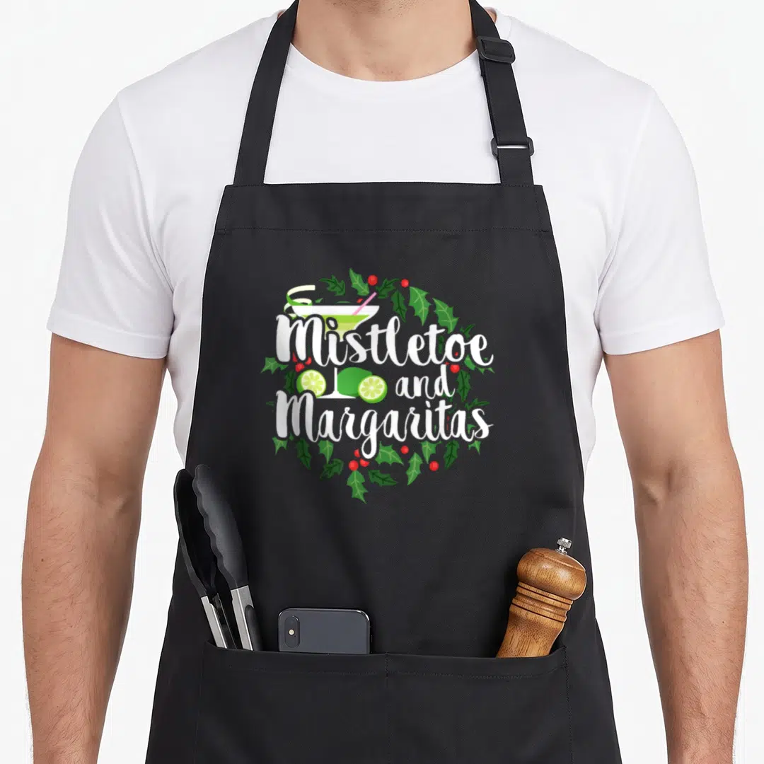Margaritas christmas apron - Mistletoe and Margaritas text with margarita glass and holly wreath in white green red — shown on a black cotton canvas apron against a white background