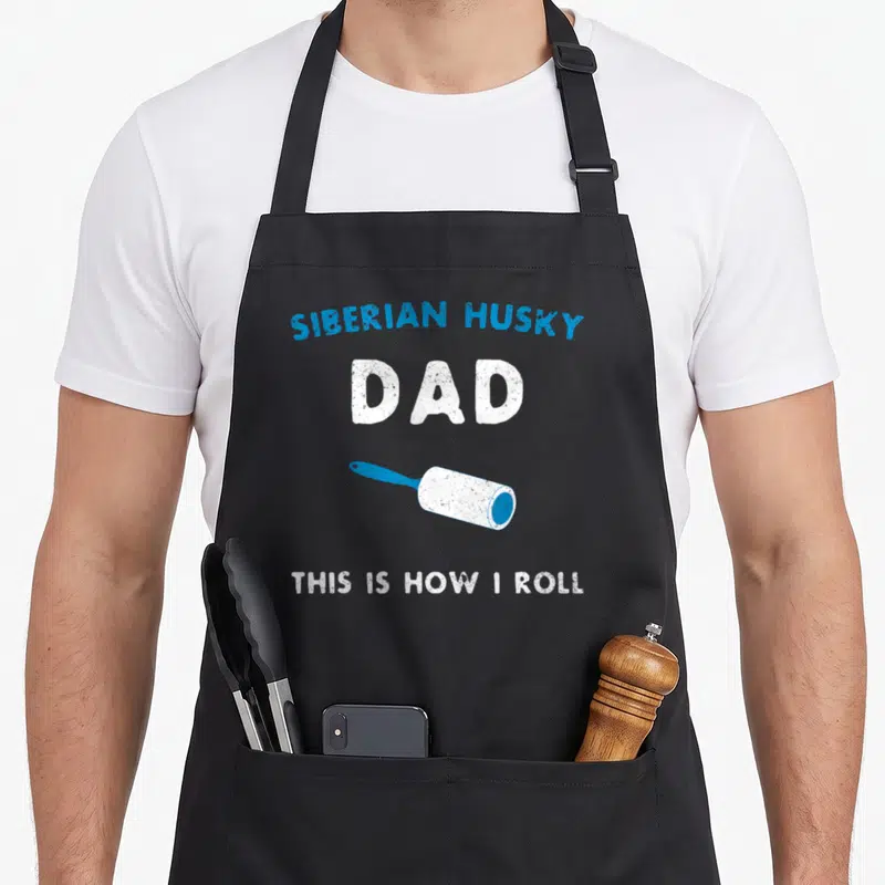 Roll dog apron - Blue and white distressed text SIBERIAN HUSKY DAD above a diagonal lint roller — shown on a black cotton canvas apron against a white background