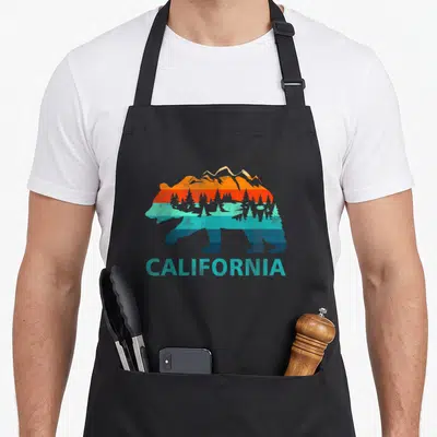 Souvenir bear apron - Retro bear silhouette with mountains trees and CALIFORNIA text in orange teal and blue — shown on a black cotton canvas apron against a white background