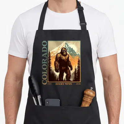 Colorado 80s apron - Bigfoot in sunglasses under mountain with vertical COLORADO text and 1876 date — shown on a black cotton canvas apron against a white background