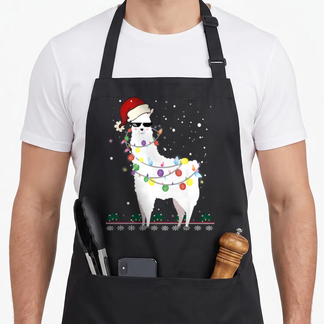 Llama xmas apron - White llama in Santa hat and sunglasses wrapped in red green blue and yellow Christmas lights above — shown on a black cotton canvas apron against a white background