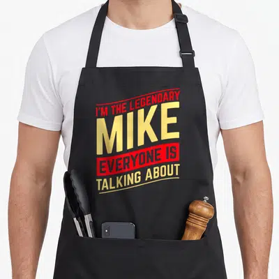 Mike christmas aprons - Red and yellow stacked text reading I'M THE LEGENDARY MIKE EVERYONE IS TALKING ABOUT — shown on a black cotton canvas apron against a white background