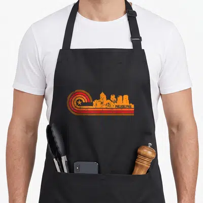 Skyline philadelphia apron - Retro orange skyline silhouette next to a concentric circle with a star and PHILADELPHIA — shown on a black cotton canvas apron against a white background