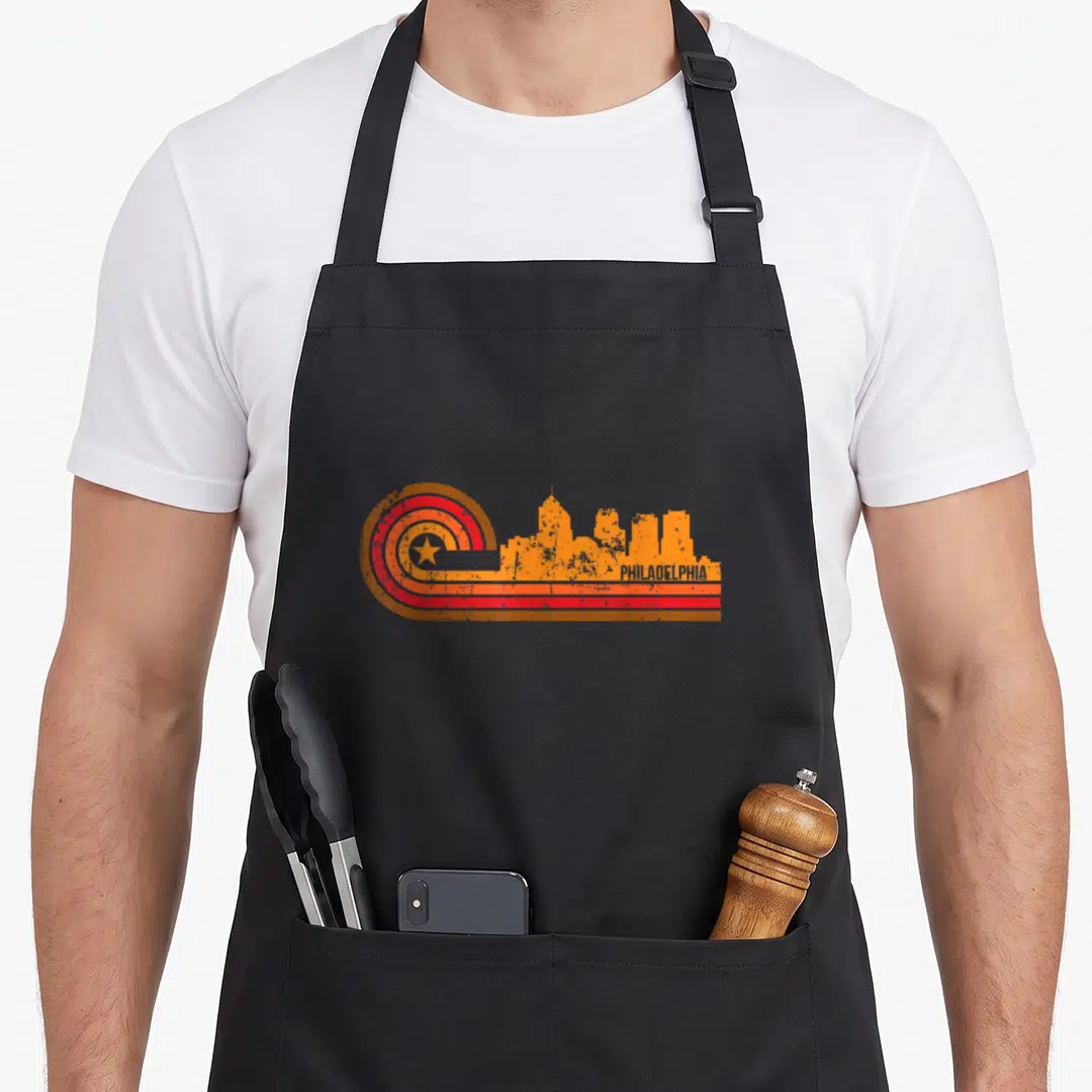 Skyline philadelphia apron - Retro orange skyline silhouette next to a concentric circle with a star and PHILADELPHIA — shown on a black cotton canvas apron against a white background
