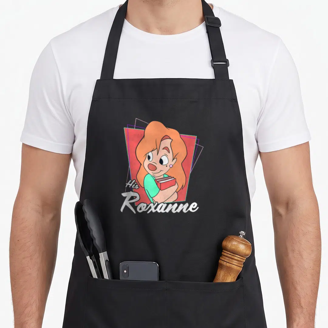 His valentine apron - Cartoon woman with orange hair holding red book next to His Roxanne text — shown on a black cotton canvas apron against a white background