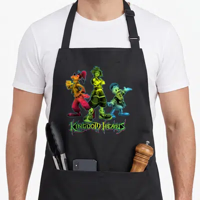 Sora duck apron - Three cartoon characters stand side-by-side above the text KINGDOM HEARTS in green and yellow — shown on a black cotton canvas apron against a white background