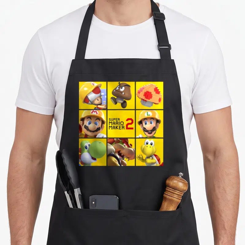 Character maker apron - 3x3 grid of Super Mario Maker 2 characters in yellow, red, blue, green with central game title — shown on a black cotton canvas apron against a white background