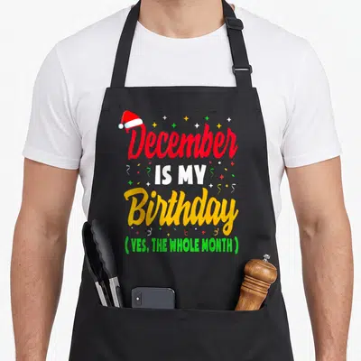 Vintage december birthday apron - Stacked December is my birthday text with santa hat and confetti in red white yellow — shown on a black cotton canvas apron against a white background