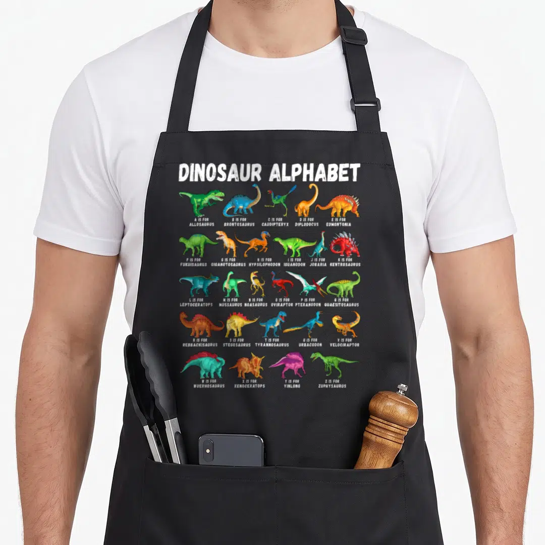 Abc dinosaur apron - Dinosaur Alphabet A-Z grid with cartoon illustrations and white labels — shown on a black cotton canvas apron against a white background