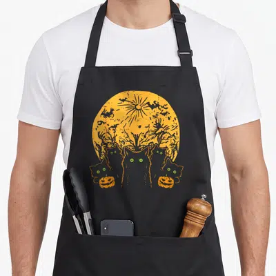 Moon halloween cat apron - Five grey cats with green eyes under an orange moon with witch silhouette and two pumpkins — shown on a black cotton canvas apron against a white background