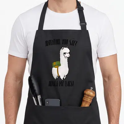 Adventure bag alpaca apron - Cartoon white alpaca with green bag, black text ADVENTURE YOU SAY? ALPACA MY BAGS! — shown on a black cotton canvas apron against a white background