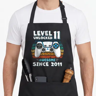 11yr bday birthday apron - Game controller with Level 11 Unlocked text and pixelated background — shown on a black cotton canvas apron against a white background