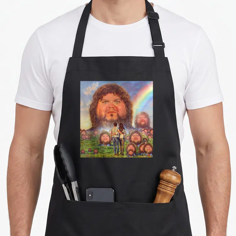 Vance meme apron - Giant bearded man head in sky above couple in field of smaller identical faces with rainbow — shown on a black cotton canvas apron against a white background