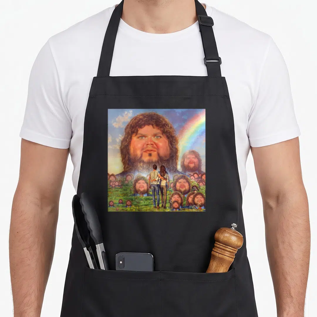 Vance meme apron - Giant bearded man head in sky above couple in field of smaller identical faces with rainbow — shown on a black cotton canvas apron against a white background