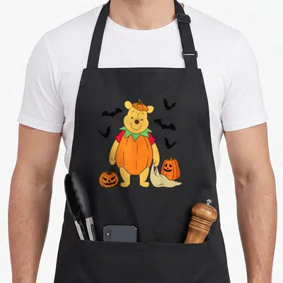 Pumpkin spooky kitchen aprons - Cartoon Winnie the Pooh in orange pumpkin costume holding bag near two jack-o'-lanterns — shown on a black cotton canvas apron against a white background