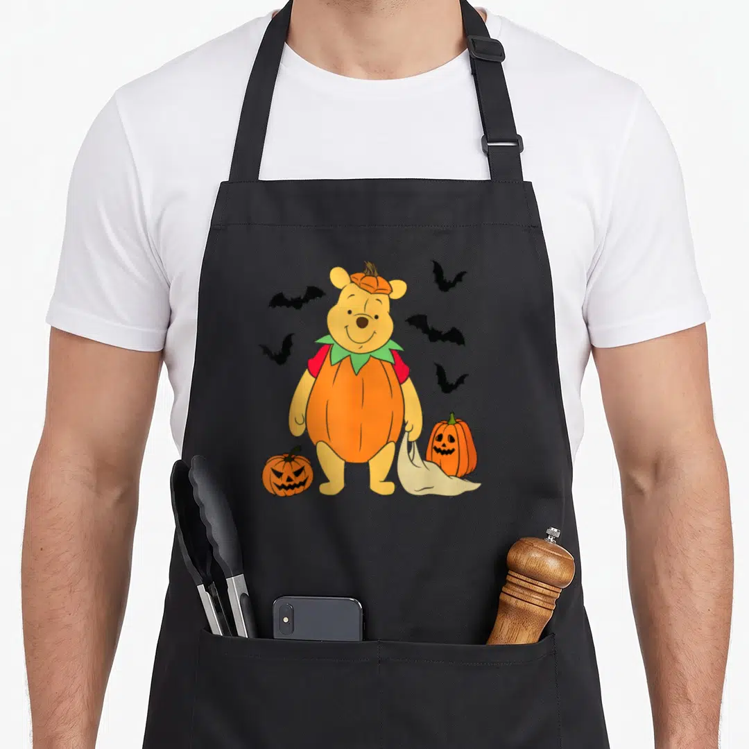 Pumpkin spooky kitchen aprons - Cartoon Winnie the Pooh in orange pumpkin costume holding bag near two jack-o'-lanterns — shown on a black cotton canvas apron against a white background