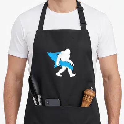 Upper bigfoot apron - White bigfoot silhouette carrying blue Upper Peninsula Michigan map shape — shown on a black cotton canvas apron against a white background