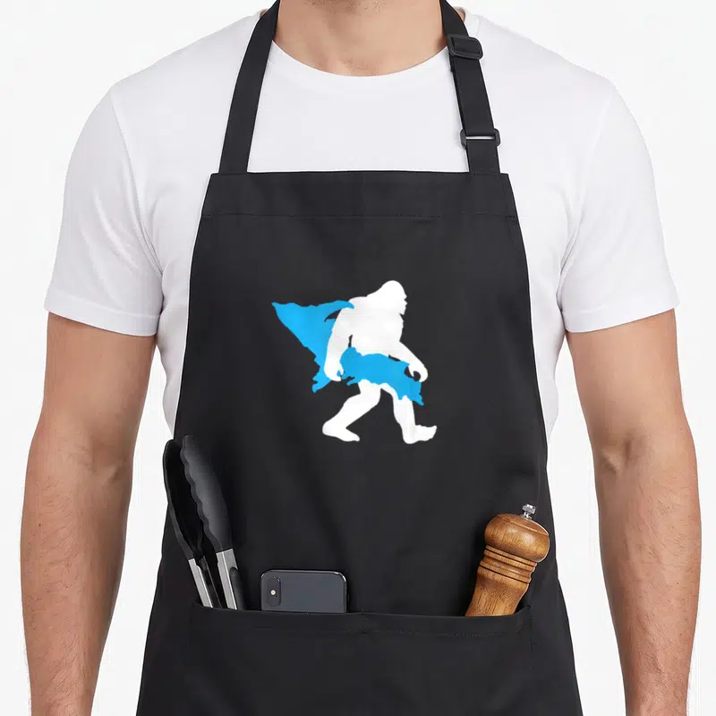 Upper bigfoot apron - White bigfoot silhouette carrying blue Upper Peninsula Michigan map shape — shown on a black cotton canvas apron against a white background