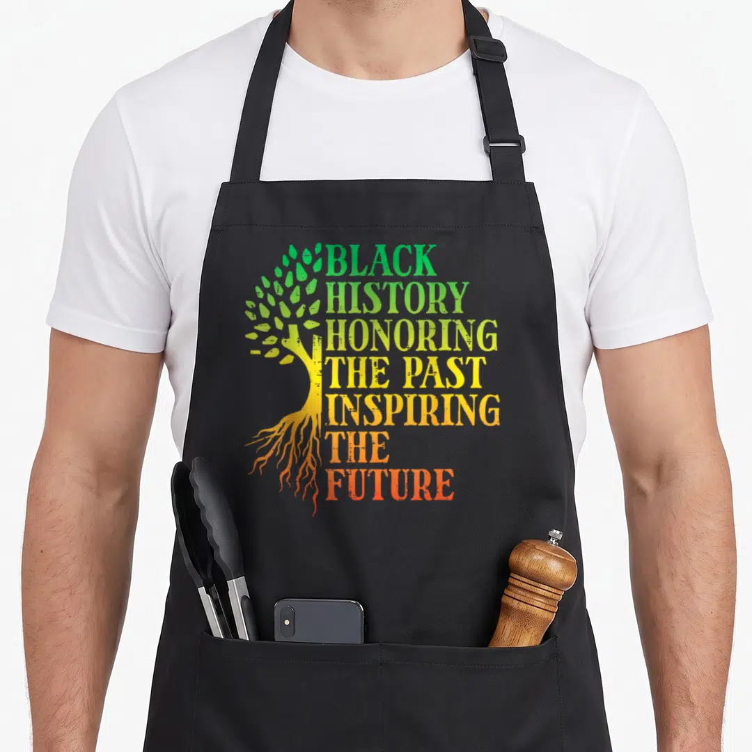 Past inspiring future history apron - Stylized tree with gradient roots and stacked text reading Black History Honoring — shown on a black cotton canvas apron against a white background