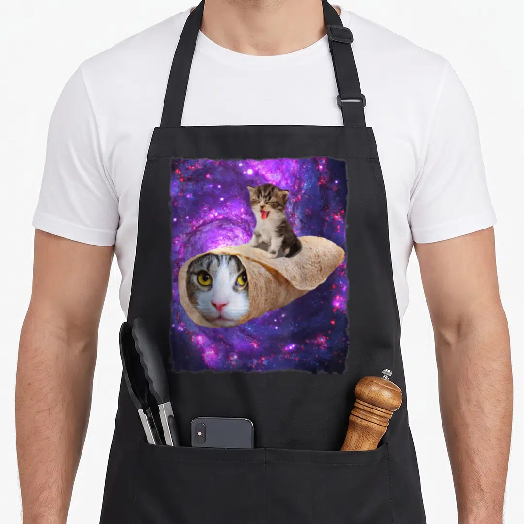 Burrito space cat apron - Cat face in tortilla with yawning kitten on top against purple blue galaxy — shown on a black cotton canvas apron against a white background