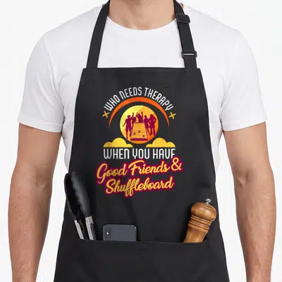 Therapy friends apron - Arched text WHO NEEDS THERAPY over yellow sun with purple shuffleboard player silhouettes — shown on a black cotton canvas apron against a white background