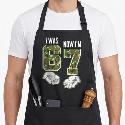 Birthday meme apron - Camo 6 and 7 numbers with I WAS NOW I'M text and two open white palms below — shown on a black cotton canvas apron against a white background