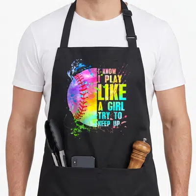 Play softball apron - Rainbow watercolor softball with stacked colorful text I KNOW I PLAY LIKE A GIRL TRY TO KEEP UP — shown on a black cotton canvas apron against a white background