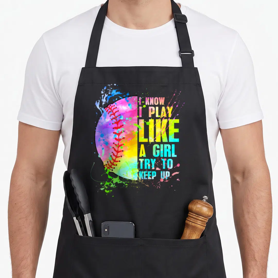 Play softball apron - Rainbow watercolor softball with stacked colorful text I KNOW I PLAY LIKE A GIRL TRY TO KEEP UP — shown on a black cotton canvas apron against a white background