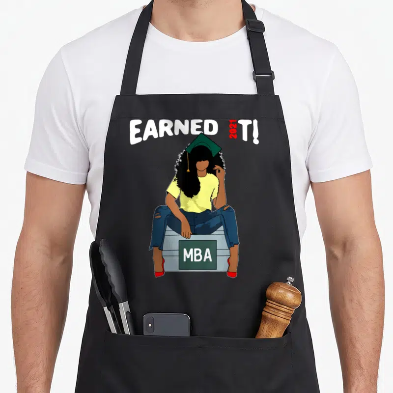 Earned master apron - Cartoon woman in cap sitting on MBA box under arched EARNED 2021 T! text — shown on a black cotton canvas apron against a white background