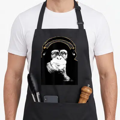 Funny monkey chimpanzee apron - Black and white photo of a chimpanzee wearing headphones resting chin on hand — shown on a black cotton canvas apron against a white background