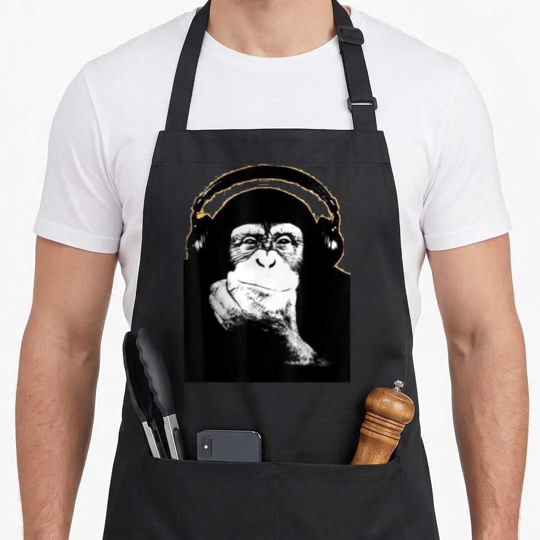 Funny monkey chimpanzee apron - Black and white photo of a chimpanzee wearing headphones resting chin on hand — shown on a black cotton canvas apron against a white background
