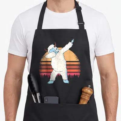 Snowman yeti apron - White yeti dabbing in front of retro orange striped sun and pink mountain silhouette — shown on a black cotton canvas apron against a white background
