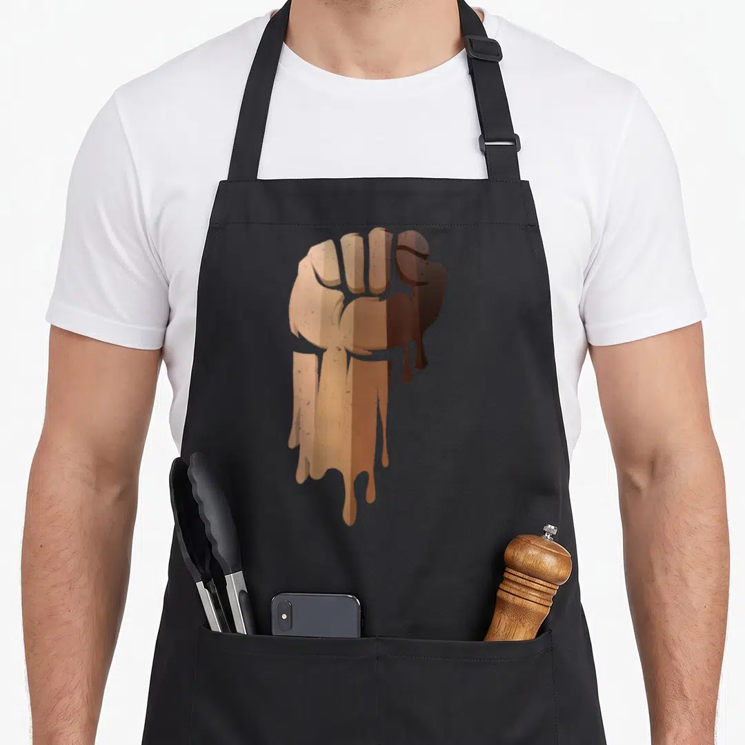 Drippin african apron - Brown and tan cartoon clenched fist with dripping effect — shown on a black cotton canvas apron against a white background