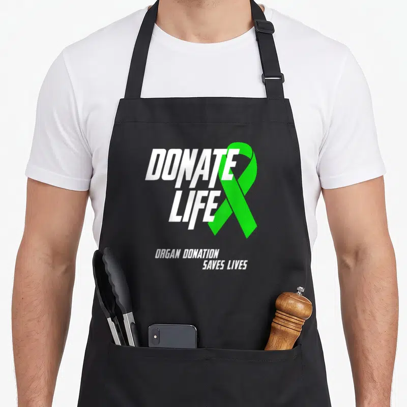 Save organ apron - White DONATE LIFE text next to green ribbon with organ donation slogan — shown on a black cotton canvas apron against a white background