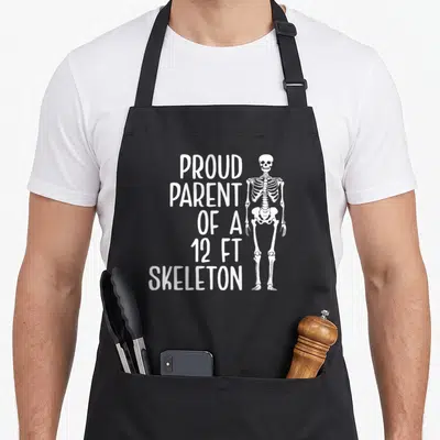 Parent skeleton apron - White stacked text reading Proud Parent of a 12ft Skeleton beside a standing cartoon skeleton — shown on a black cotton canvas apron against a white background