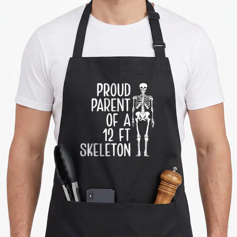 Parent skeleton apron - White stacked text reading Proud Parent of a 12ft Skeleton beside a standing cartoon skeleton — shown on a black cotton canvas apron against a white background