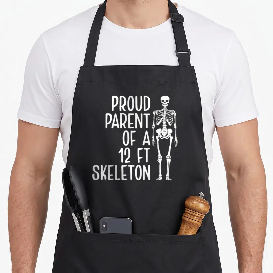 Parent skeleton apron - White stacked text reading Proud Parent of a 12ft Skeleton beside a standing cartoon skeleton — shown on a black cotton canvas apron against a white background