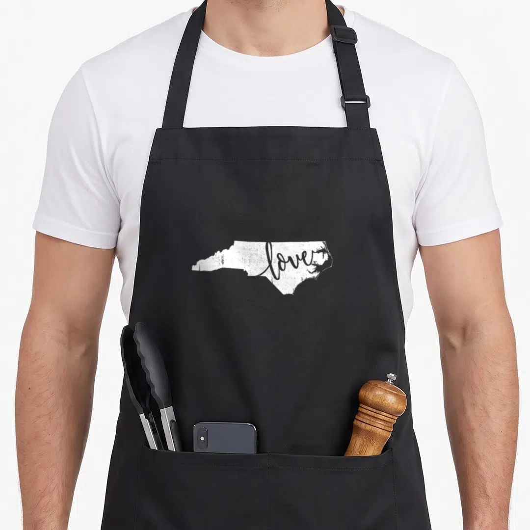 North carolina pride apron - White North Carolina state silhouette with love script overlay in distressed style — shown on a black cotton canvas apron against a white background