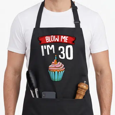Blow cake apron - Red ribbon with BLOW ME I'M 30 text above a pink frosted cupcake with a blue candle — shown on a black cotton canvas apron against a white background