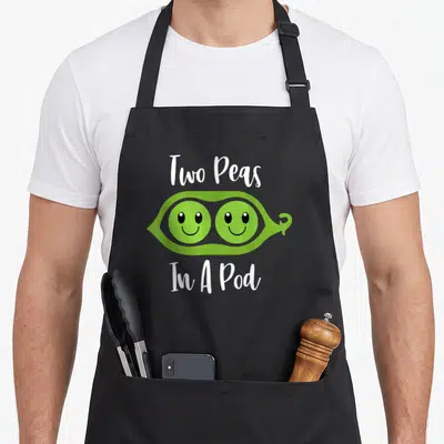 Peas couple apron - Green cartoon pea pod with two smiling faces and white text Two Peas In A Pod — shown on a black cotton canvas apron against a white background