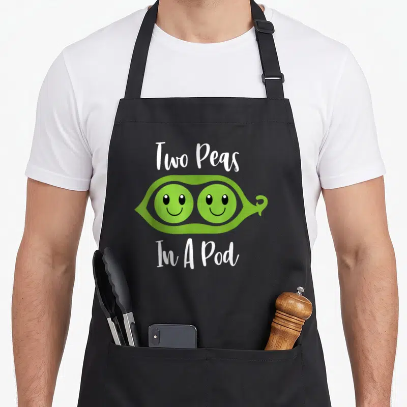 Peas couple apron - Green cartoon pea pod with two smiling faces and white text Two Peas In A Pod — shown on a black cotton canvas apron against a white background