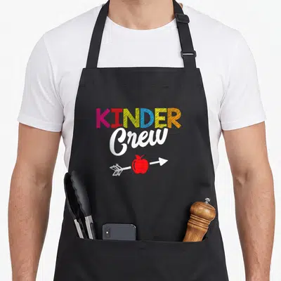 Crew student kindergarten teacher apron - KINDER Crew text with pink red yellow blue orange letters and red apple — shown on a black cotton canvas apron against a white background