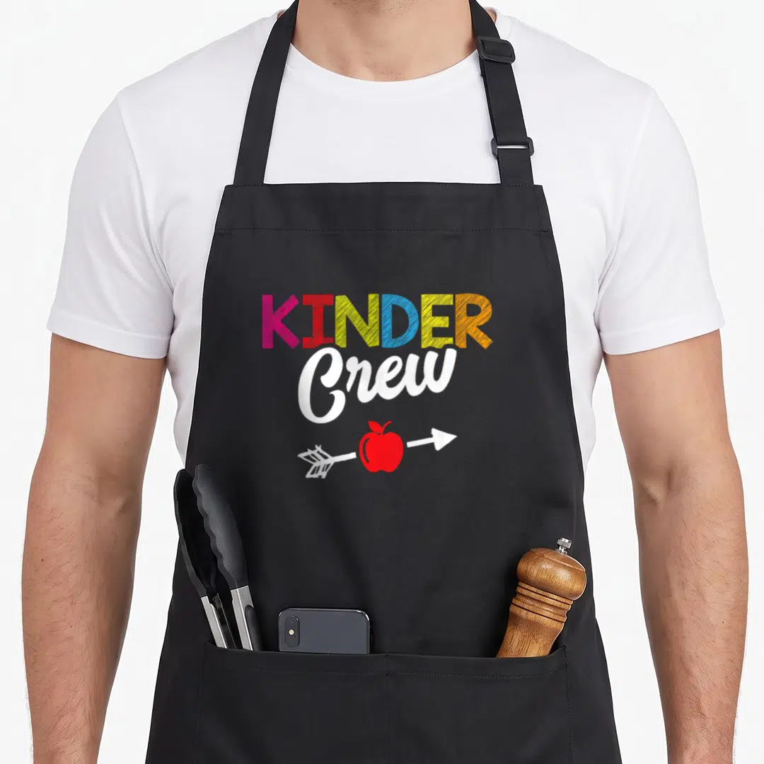 Crew student kindergarten teacher apron - KINDER Crew text with pink red yellow blue orange letters and red apple — shown on a black cotton canvas apron against a white background